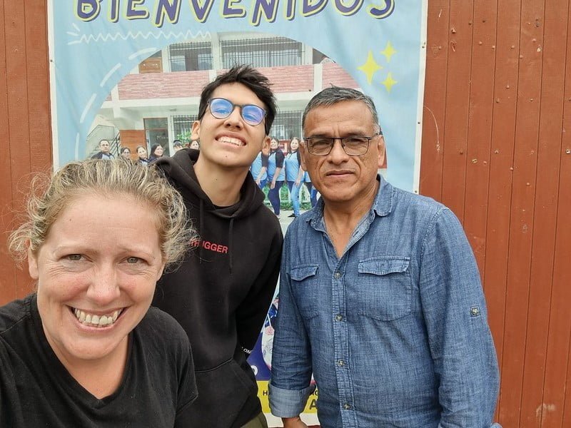 One woman and 2 men in front of a poster for a school. Writing on the poster says 'Beinvenidos'